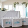 Three personalised glossy white gift bags are arranged against a wedding-themed background dominated by light blue and white colors. The bags have white rope handles, and one bag, positioned centrally, is clearly labeled with the name "Nikki" in a flowing blue script font. The surrounding decor includes lush light blue and white floral arrangements and possibly some greenery, setting a sophisticated and romantic wedding party mood.