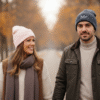 A close-up, face-on lifestyle image of a couple and two small children sitting on a snowy bench outdoors, smiling and wearing matching white bobble hats. The woman's hat has "Mrs" embroidered on the front, and the man's hat has "Mr" embroidered on the front. They are all bundled up in warm clothing, with a snowy winter landscape and pine trees visible in the soft background.