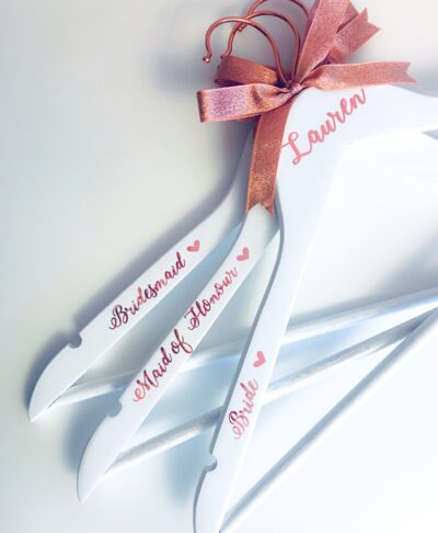 Personalised white bridal party hangers with rose gold hooks and bows, featuring custom names and titles such as Bride, Bridesmaid, and Maid of Honour — elegant customised wedding morning accessories