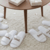 lifestyle shot of several pairs of white open-toe spa slippers arranged on a plush cream carpet. Each slipper features personalised names like 'Sarah' and 'Melinda' or titles like 'Maid of Honour' in elegant rose gold cursive script. A wooden coffee table with white gift boxes and pink confetti is partially visible in the blurred background.