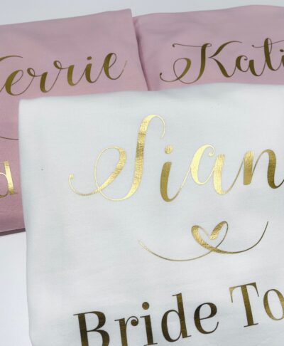 Close-up of three folded personalised hoodies: two light pink and one white. The white hoodie is in the foreground and features the name 'Sian' in large, metallic gold script, with 'Bride To Be' underneath. The pink hoodies in the background have names like 'Kerrie' and 'Katie' printed in gold script, with 'Bridesmaid' underneath.