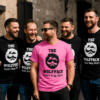 A group of six men are standing together outdoors, laughing and smiling while wearing matching black t-shirts that read “THE WOLFPACK Kyle’s Stag 2024.” One man in the center stands out in a bright pink version of the same shirt. The group appears to be enjoying a stag party on a sunny day, with stone buildings in the background.