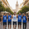 Five men are walking toward the camera on a cobblestone street in a historic European city, likely Seville, Spain. They are all wearing tank tops with a "GAME OVER Chris's Stag Spain 2024" graphic. Four men wear blue vests with white text, and the man in the center wears a white vest with blue text. The men on the ends are holding a glass bottle of beer. The background features yellow buildings with orange trees and the Giralda bell tower in the distance.