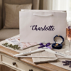 A personalised wooden bridesmaid hanger and gift box set on an elegant white table, featuring a custom name "Charlotte", an amethyst purple ribbon, and eucalyptus and lavender accents.