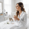 A bride is sitting and smiling in a beautifully lit room on the morning of her wedding, wearing long sleeved white satin pyjamas personalised with black text, and holding a glass of champagne.