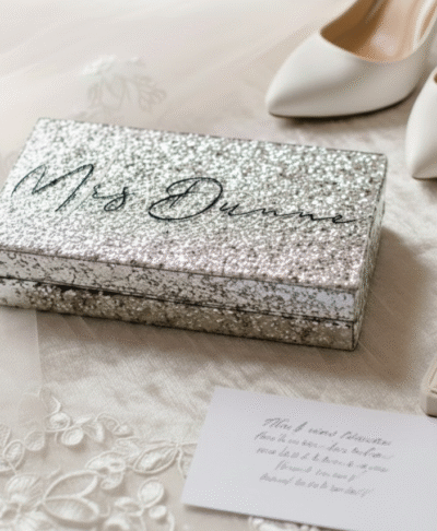 A silver glitter clutch bag is displayed on white, lace edged bridal fabric next to a pair of white pointed toe high heeled shoes, an open beige ring box containing a diamond engagement ring, and a small handwritten note on a white card.