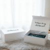 A lifestyle photo of a white bridesmaid gift box open on a furry rug. Inside are green satin items labeled 'Rebecca Bridesmaid' and a hand placing a personalised champagne flute inside. A second box lid labeled 'Will you be my bridesmaid?' and a third lid labeled 'Hanna' are visible. White roses are in the background.