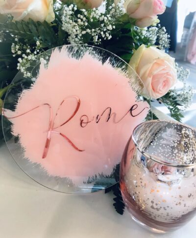 Clear acrylic circle table sign with pink painted background and rose gold lettering, styled with flowers and candles for wedding table décor.
