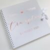 Personalised 12×12 wedding guest book with names, date, and surname in metallic script on a white cover with satin ribbon tie.