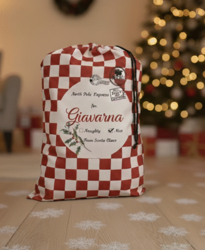 Luxury personalised Santa sacks for kids, featuring a red and white checkered North Pole Express bag with the name 'Giavarna'. Perfect Christmas morning setting on a hardwood floor with snow flake details, blurred Christmas tree lights, and cosy home decor.