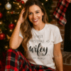 A smiling woman sits on the floor beside a decorated Christmas tree, wearing a white T-shirt that says “First Christmas as wifey, Est. 06.08.21” and red and black plaid pajama pants. The cozy setting includes a lit fireplace, stockings, and warm festive lighting.