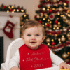 A close-up image of a baby wearing a red bib with a festive print. The background is softly blurred but has warm, cosy colors suggestive of a Christmas setting.