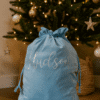 A light blue velvet Christmas sack with the name “Hudson” written in silver cursive sits on a wooden floor in front of a decorated Christmas tree adorned with gold and white ornaments and warm fairy lights.