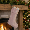 Luxury personalised pink velvet Christmas stocking with "Olivia" in silver script, filled with small gifts and hanging on a rustic wooden mantelpiece above a cosy fireplace. Festive holiday decor with a decorated Christmas tree in the background, perfect for custom Christmas gifts and home decoration.