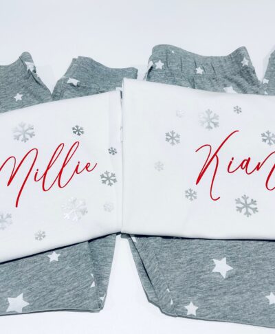These are two pairs of personalised Christmas pyjamas, featuring grey bottoms printed with white stars and matching white tops. The tops are customised with names written in a festive red script font: 
