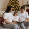 A cosy, festive Christmas lifestyle image showing a mother, father, and daughter sitting together on a large, comfortable sofa in a living room decorated for the holidays. They are all wearing matching short-sleeved white t-shirts and full-length grey pyjama trousers with a white star pattern. The background features a beautifully lit Christmas tree and a fireplace with a lit fire. The family is smiling and enjoying a moment together.