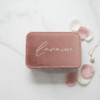 a small, rectangular travel jewellery case, closed and made of rose quartz velvet fabric. The lid is personalised with the name "Laraine" written in silver script. The case is resting on a white marble surface and is surrounded by a few scattered pink and white flower petals and a gold necklace with a pearl pendant.