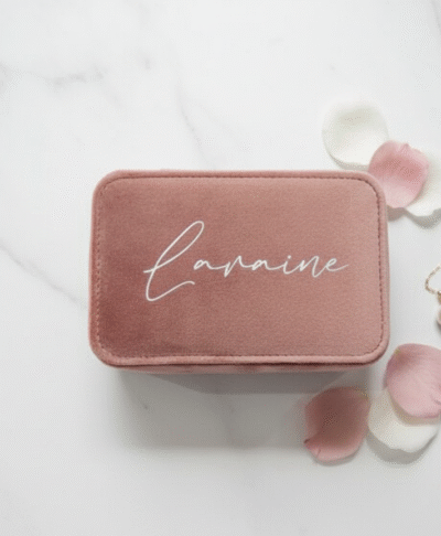 a small, rectangular travel jewellery case, closed and made of rose quartz velvet fabric. The lid is personalised with the name "Laraine" written in silver script. The case is resting on a white marble surface and is surrounded by a few scattered pink and white flower petals and a gold necklace with a pearl pendant.