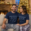 Lifestyle image of a couple wearing matching Christmas pyjama sets, with white short-sleeve t-shirts and festive check pyjama trousers. The man and woman are posing together in a cosy holiday setting with a fireplace and a decorated Christmas tree. Ideal matching couple pyjamas for Christmas morning photos.