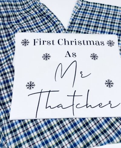 A personalised christmas pyjama set featuring check bottoms in shades of blue, green, and white, with a white. The white fabric is printed with black text and snowflake graphics that read: 