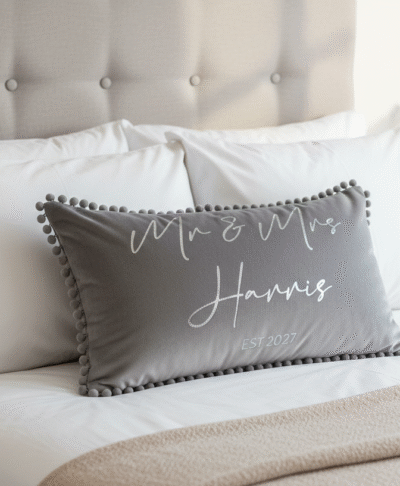 Grey personalised ‘Mr & Mrs Harris’ decorative cushion with pom-pom trim displayed on a neatly made bed with white pillows and neutral bedroom decor.