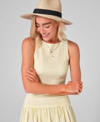 A woman smiles and looks down in a studio shot, modeling a customisable straw fedora. The natural-toned hat features a classic black ribbon band, designed to be personalised with initials for a unique bridal party gift or honeymoon accessory. She is wearing a yellow and white gingham smocked dress, silver jewellery, and a soft pink manicure, creating an ideal look for a summer wedding guest or bachelorette weekend.